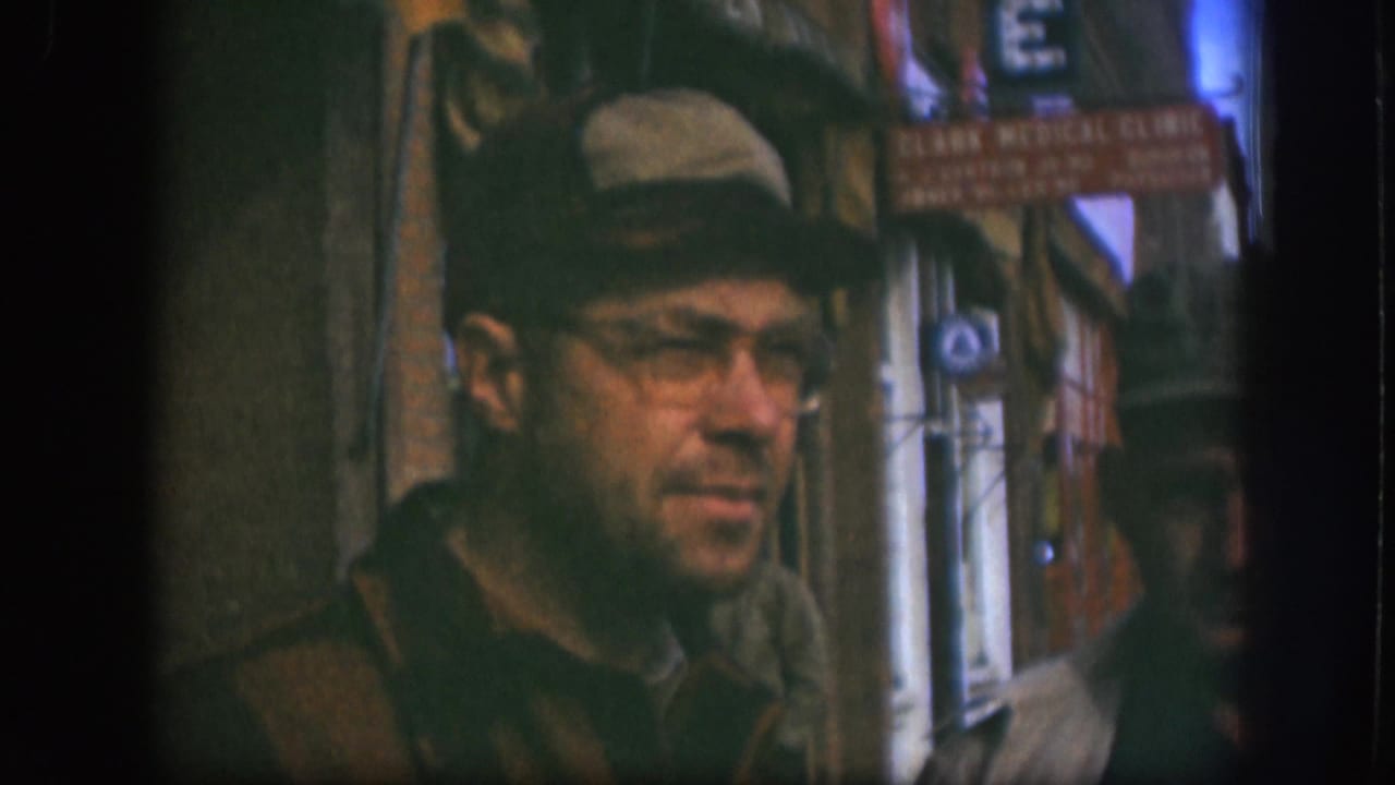 Man in a tan cap and glasses stands on a city sidewalk near storefront signage.