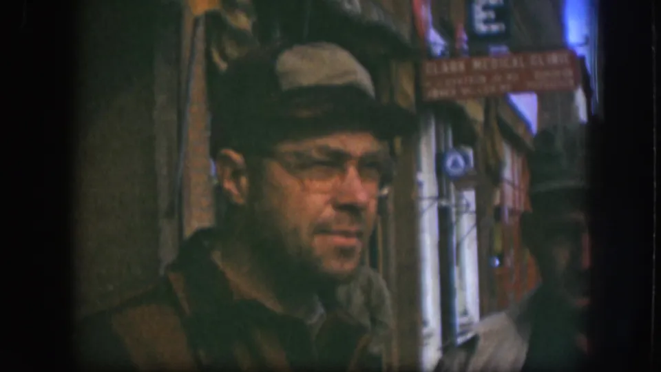 In Kansas 1958 home-movie footage, a man in a tan cap and glasses looks sideways near storefronts and a clinic sign.