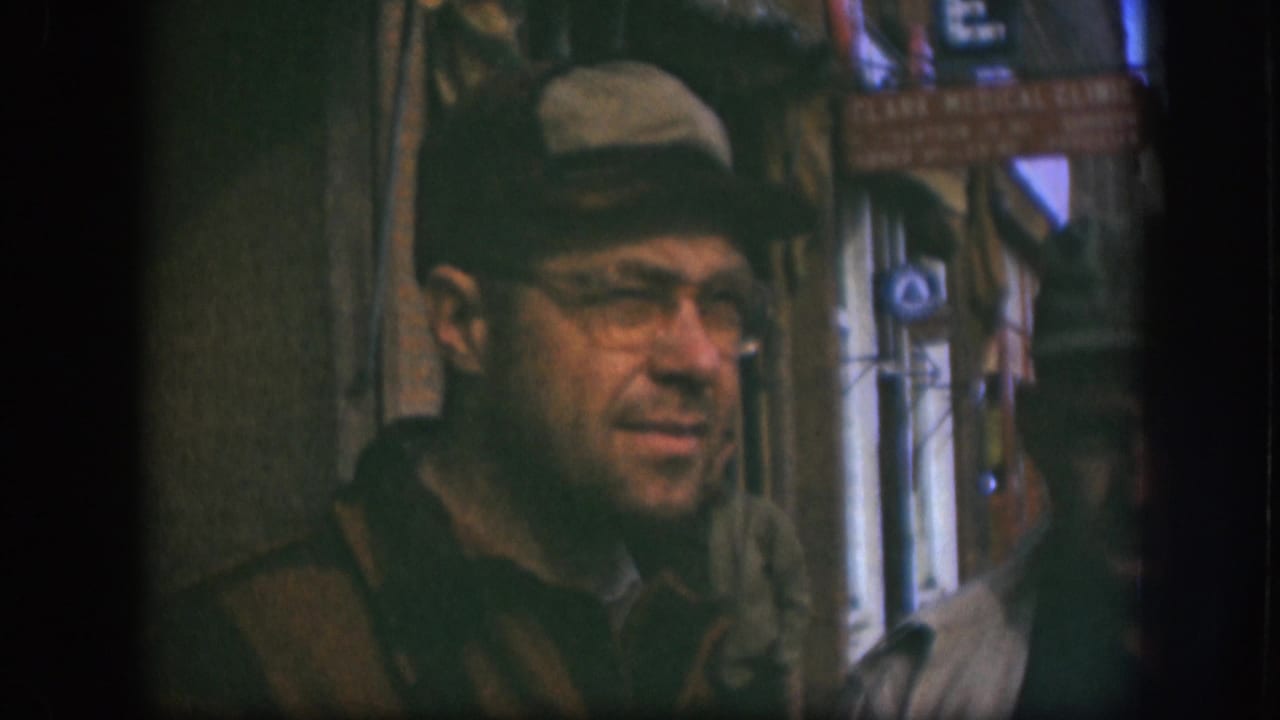 The man in a tan cap and glasses turns near the camera on a Kansas street, with storefront signs and passersby behind him.