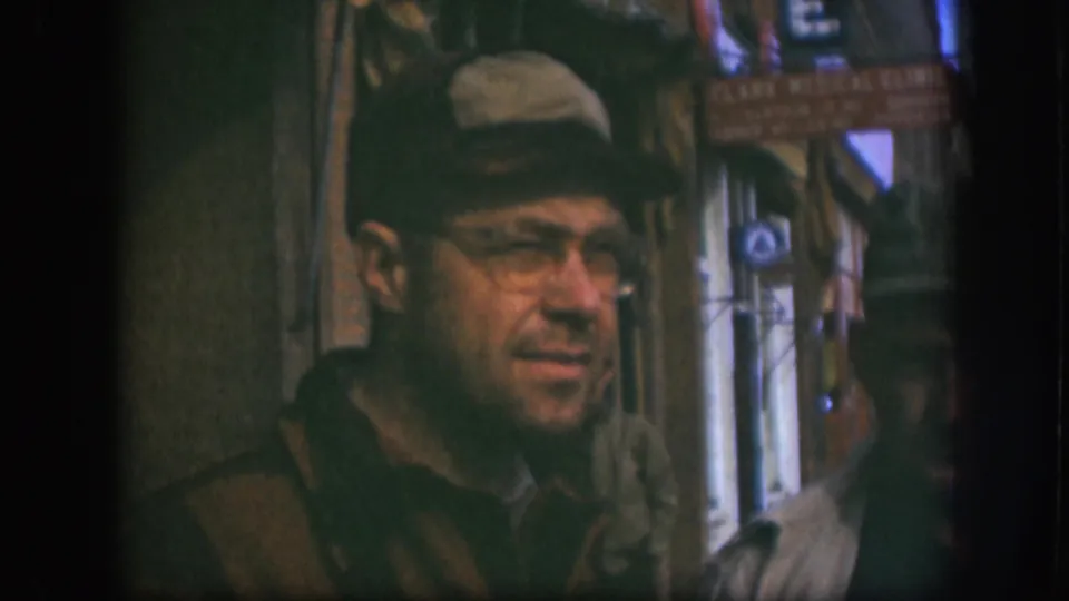 The man in a tan cap and glasses turns near the camera on a Kansas street, with storefront signs and passersby behind him.