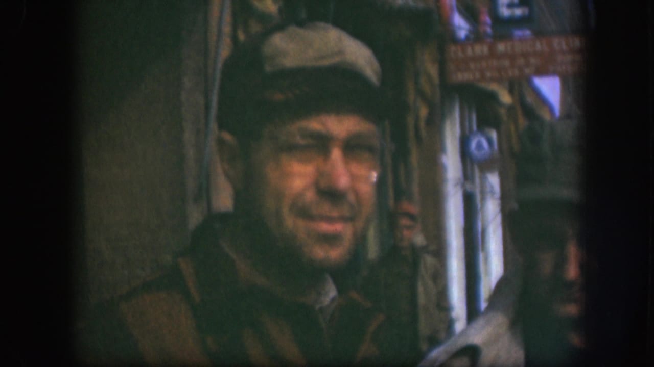 In a soft 8mm frame, the man in a tan cap and glasses looks toward the camera as a passerby moves along the Kansas sidewalk.