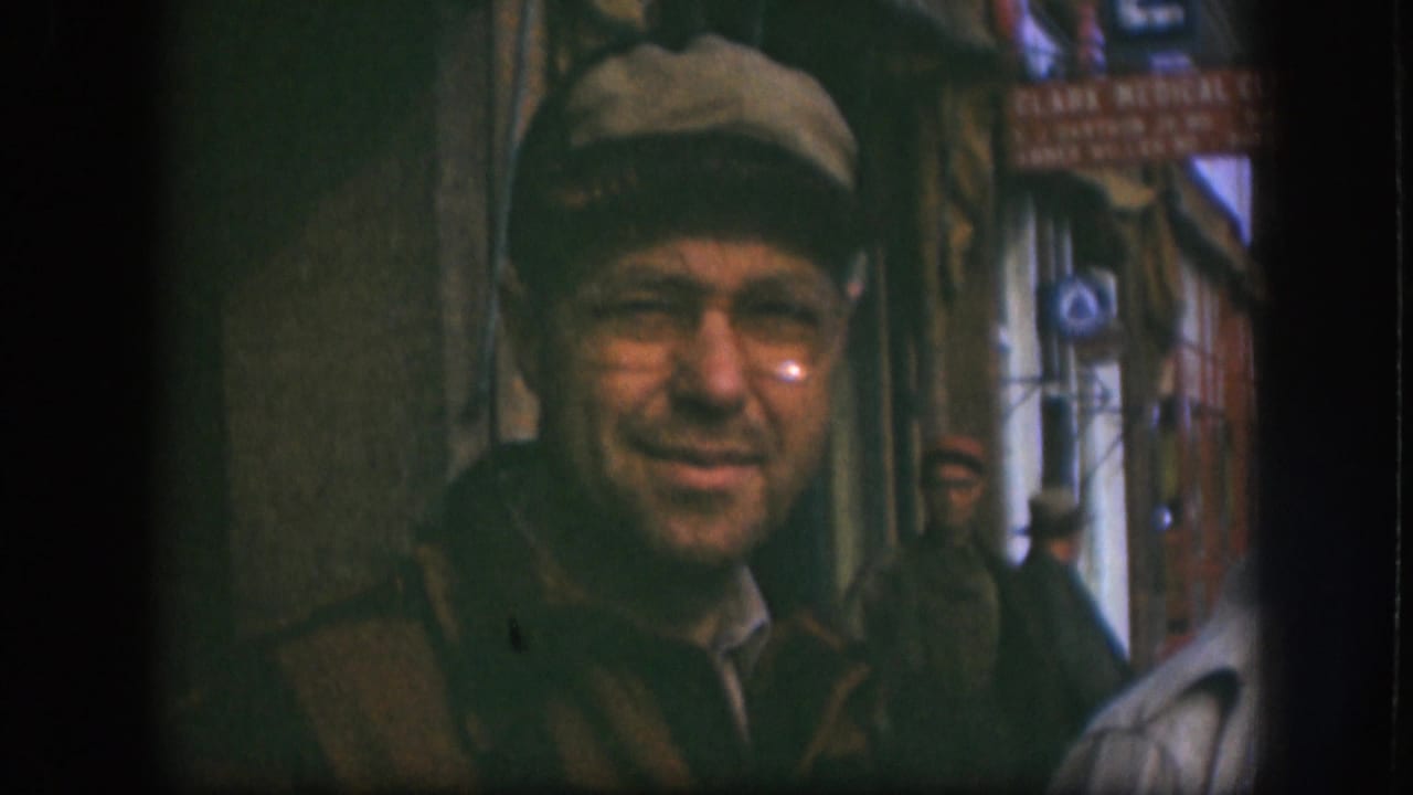 The man in a tan cap and glasses gives a slight smile toward the camera, with Kansas storefront signage and pedestrians behind him.