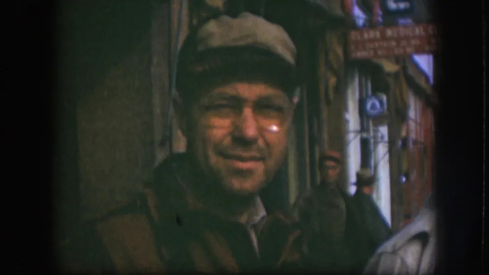 The man in a tan cap and glasses gives a slight smile toward the camera, with Kansas storefront signage and pedestrians behind him.