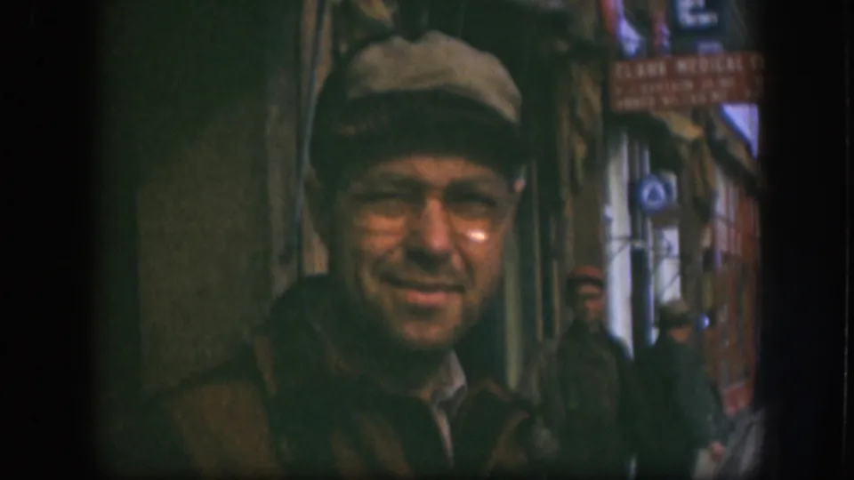 In 1958 Kansas home-movie footage, a man in a cap and glasses faces the camera while two men stand farther back in a narrow storefront passage.