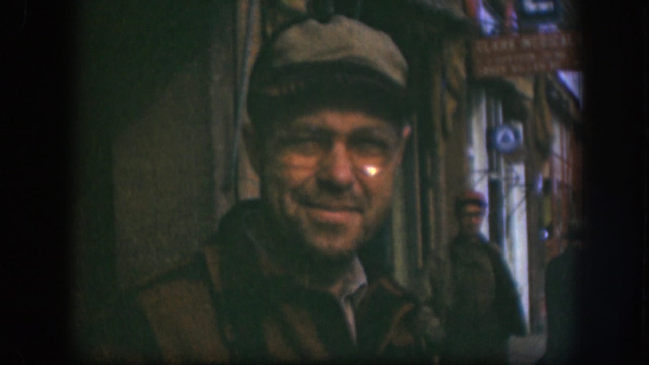 The man remains centered in soft 8mm focus, with a visible sign and two background men in the Kansas street passage.
