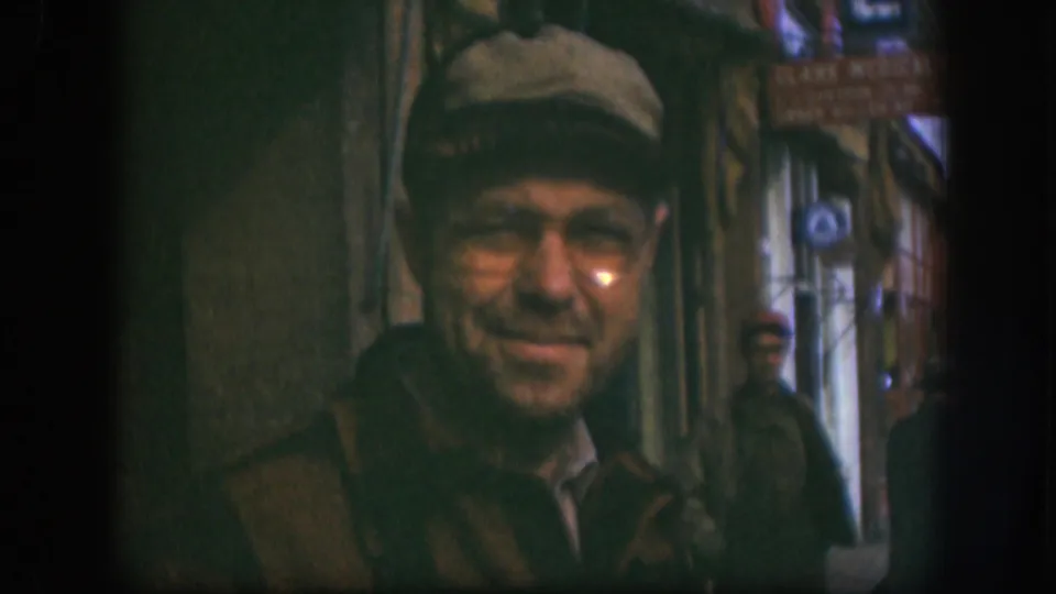 The man remains centered in soft 8mm focus, with a visible sign and two background men in the Kansas street passage.