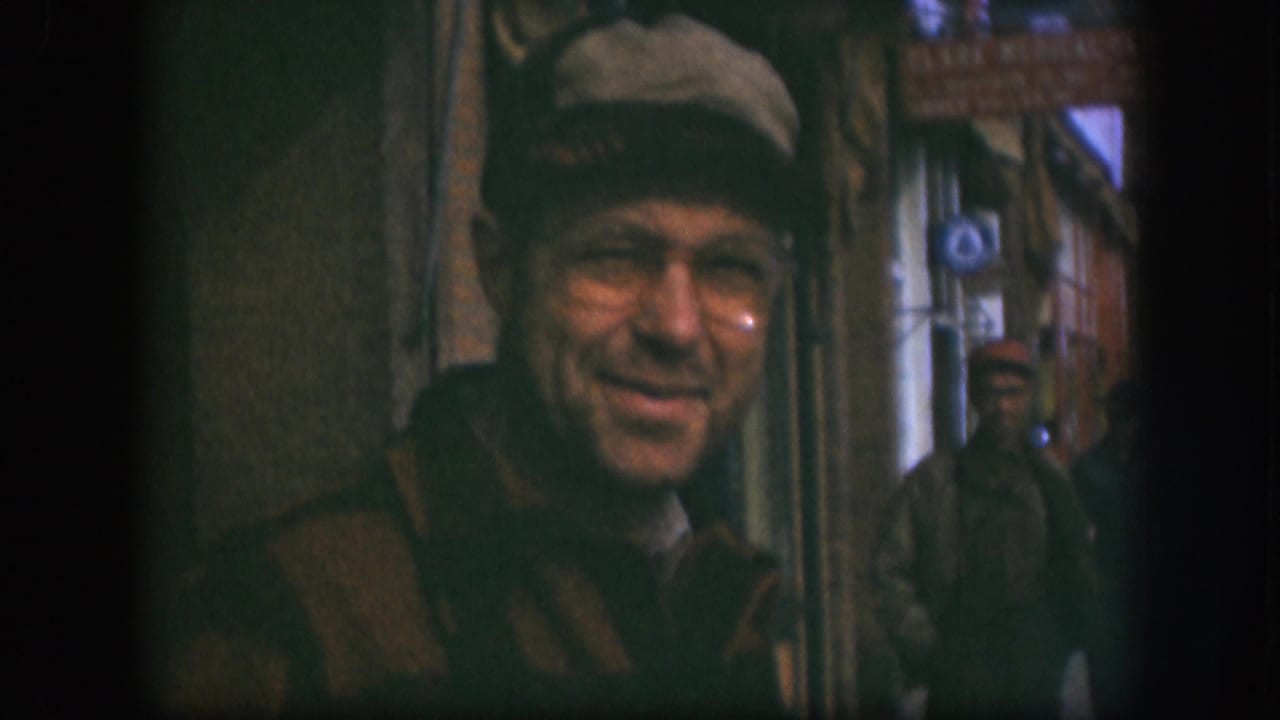 In this 1958 Kansas home-movie frame, a man in a brown cap and glasses smiles close to the camera on a sidewalk street scene.