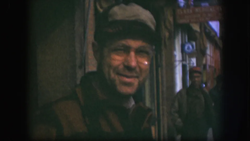 In this 1958 Kansas home-movie frame, a man in a brown cap and glasses smiles close to the camera on a sidewalk street scene.