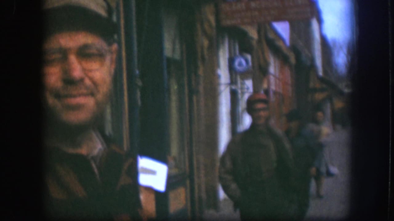 In the Kansas 1958 home-movie frame, the camera shifts so the smiling man is cropped at left and pedestrians line the storefront sidewalk.