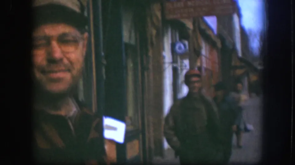 In the Kansas 1958 home-movie frame, the camera shifts so the smiling man is cropped at left and pedestrians line the storefront sidewalk.