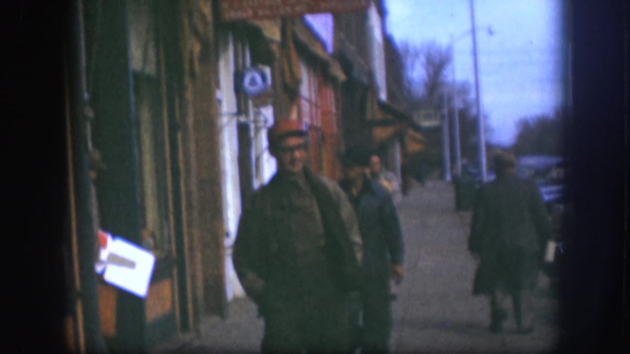 Two men walk along a sidewalk beside storefronts and parked cars.