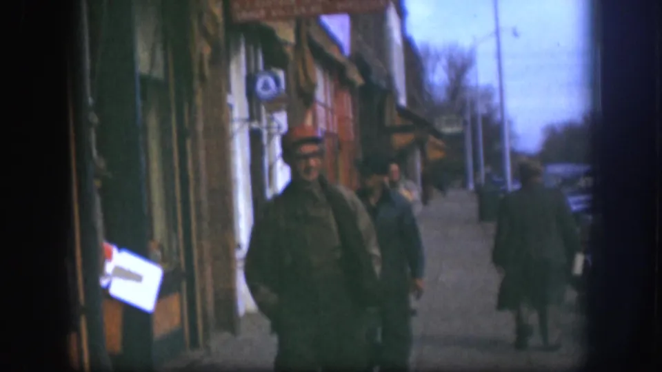 1958 Kansas 8mm frame showing two men in grey clothing walking past storefronts, with cars and pedestrians farther down the sidewalk.