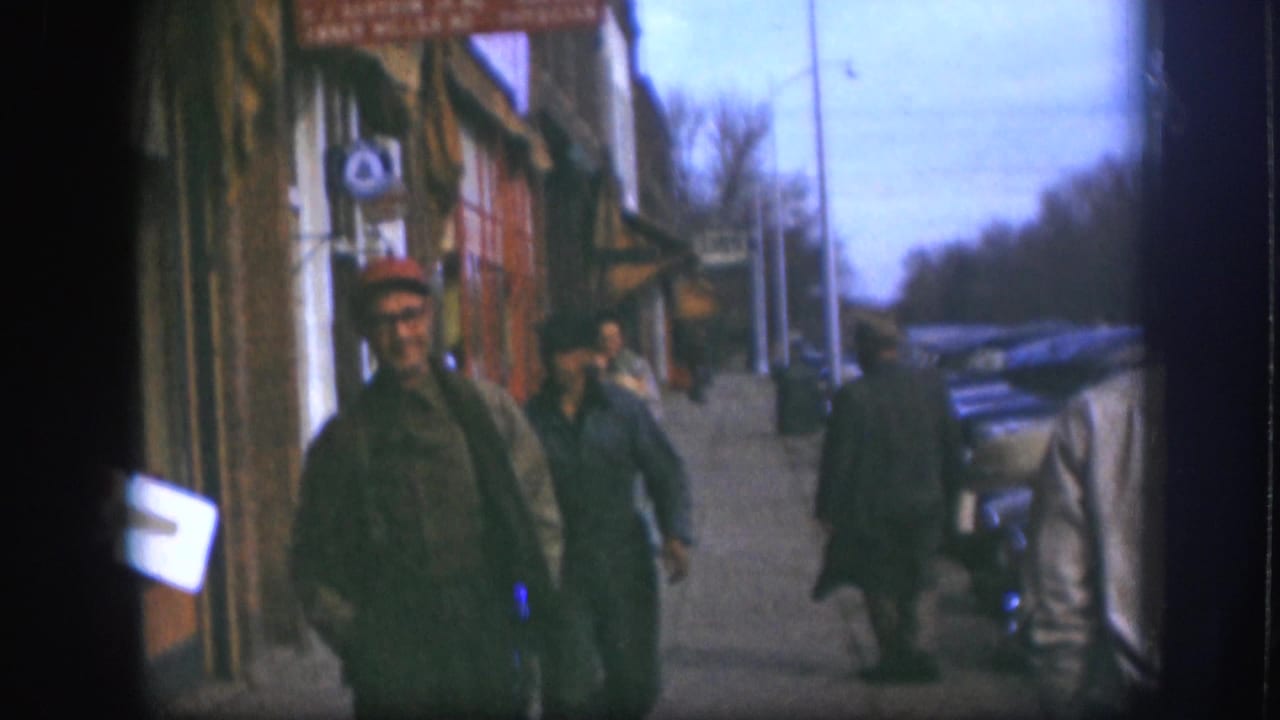 1958 Kansas street frame with men walking near storefronts, a sidewalk sign, parked cars, and bare trees in the distance.