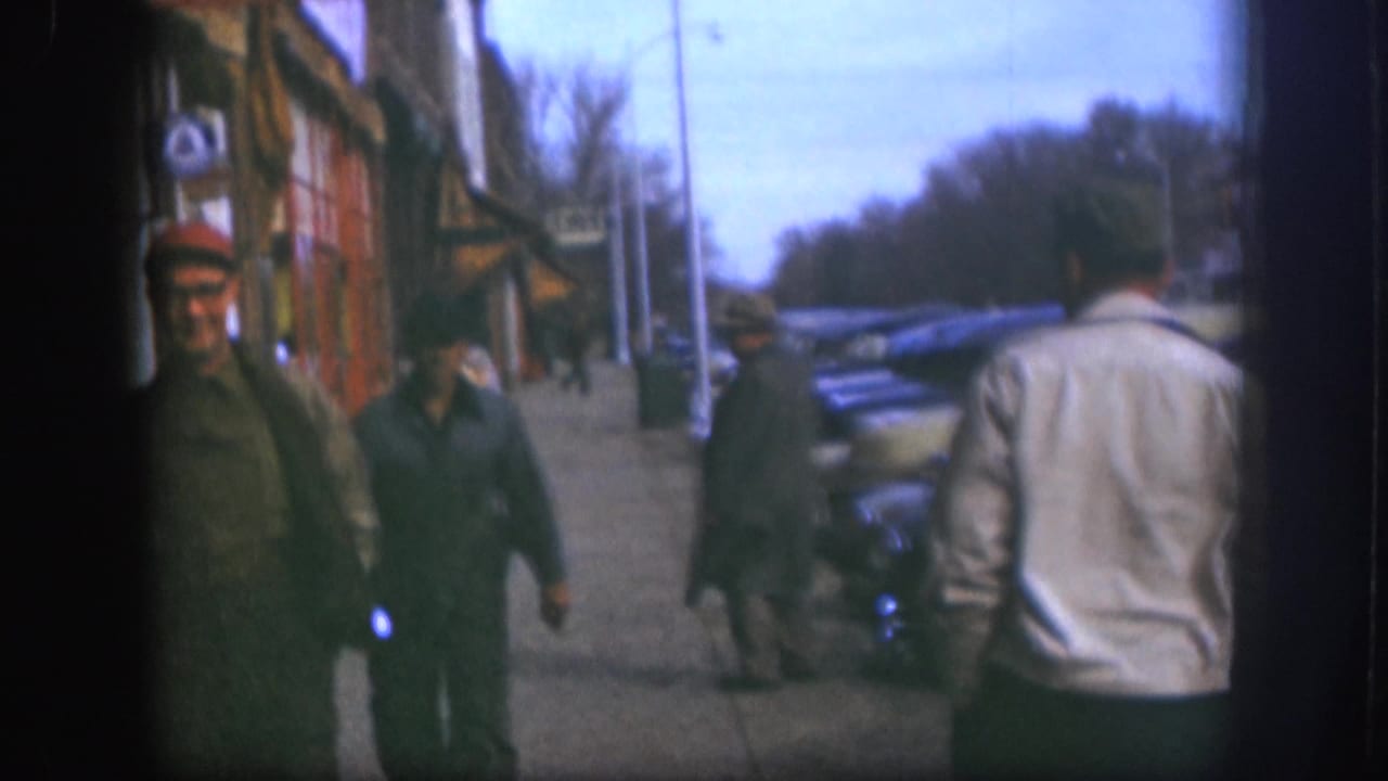 1958 Kansas sidewalk view with a man in a light jacket seen from behind, two men nearby, storefronts, and parked cars along the curb.