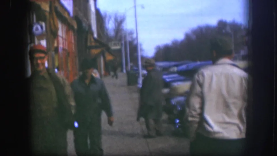 1958 Kansas sidewalk view with a man in a light jacket seen from behind, two men nearby, storefronts, and parked cars along the curb.