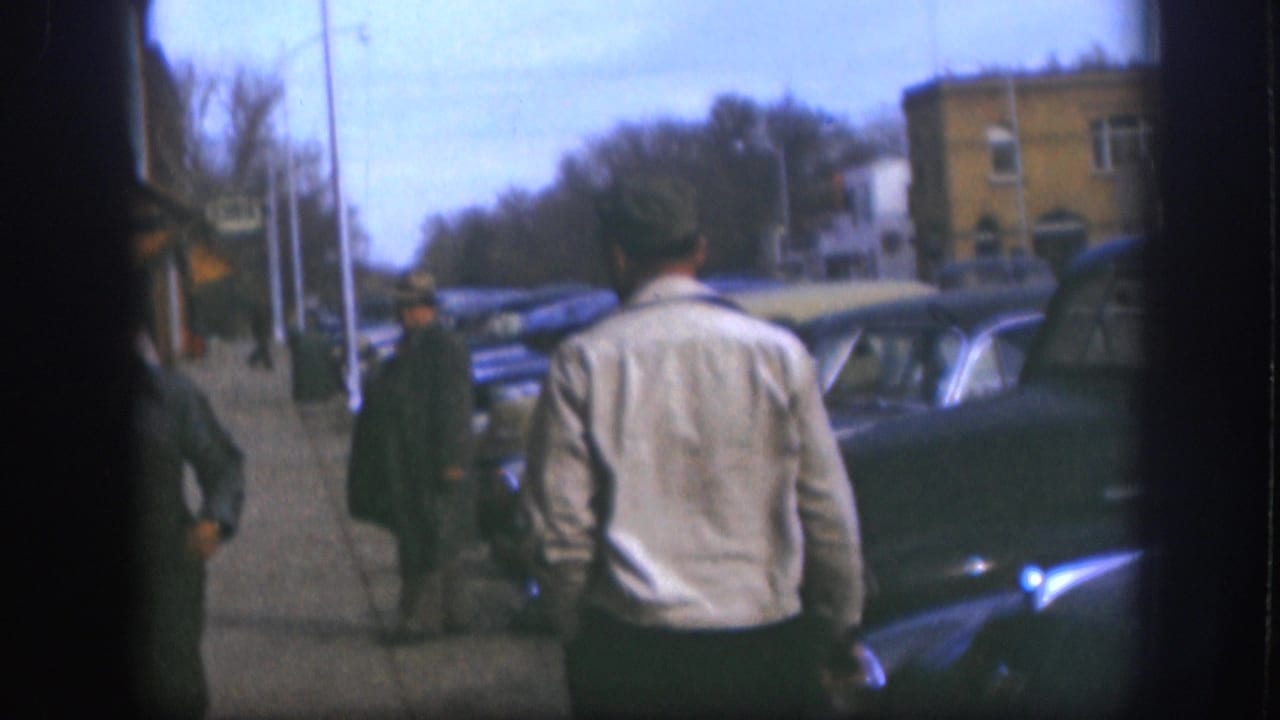 1958 Kansas 8mm frame showing a man in a light jacket from behind, with pedestrians, storefronts, parked cars, and bare trees ahead.