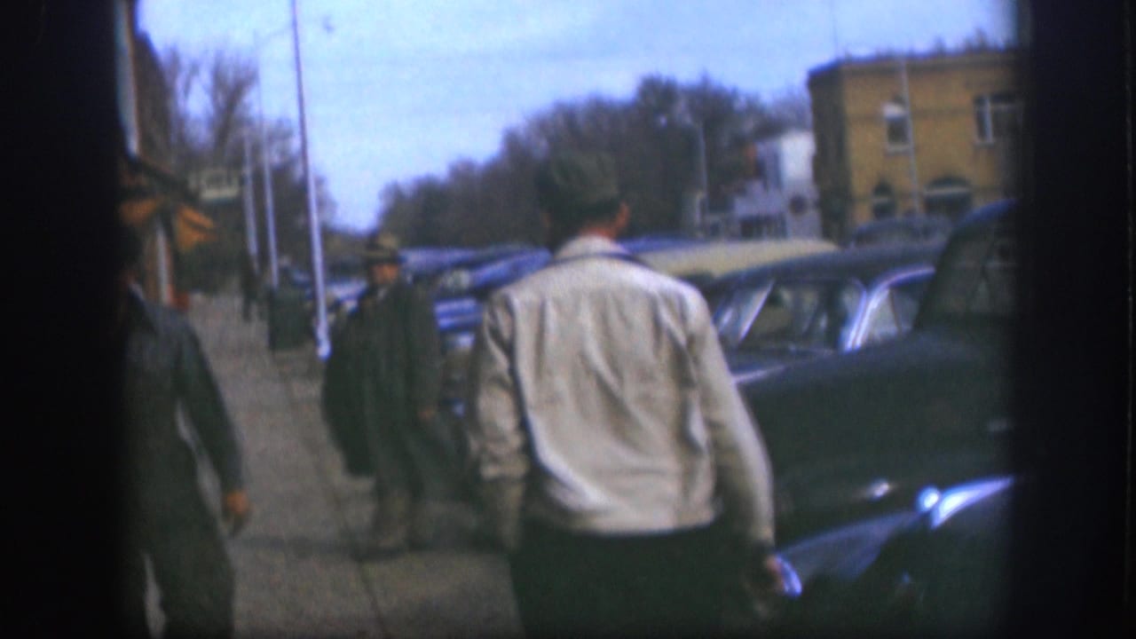 1958 Kansas 8mm frame showing men walking on a sidewalk beside parked vintage cars, storefronts, and bare trees.