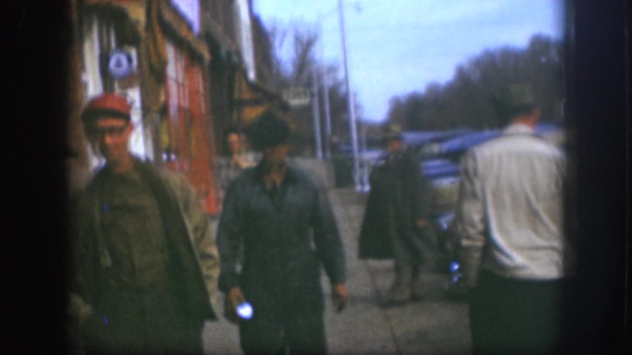 1958 Kansas street frame with men in work clothes and caps passing storefronts as a man in a white sweater walks away.
