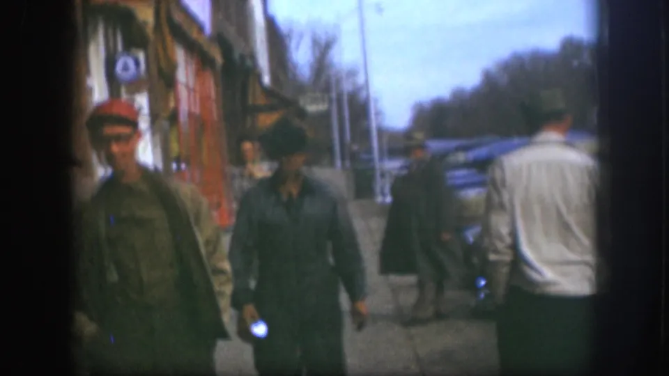 1958 Kansas street frame with men in work clothes and caps passing storefronts as a man in a white sweater walks away.
