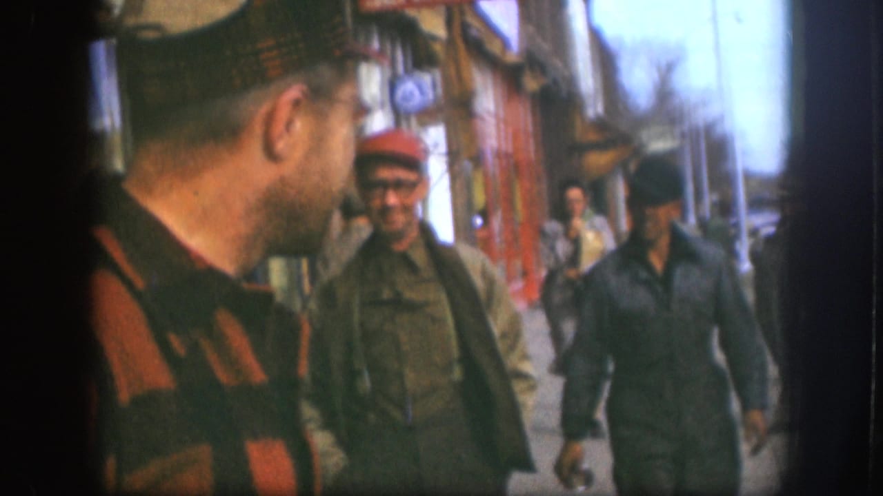 1958 Kansas sidewalk view with a striped-shirted man in the foreground and two men in caps approaching near storefronts.