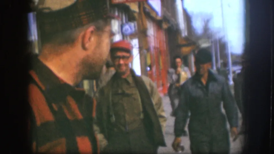 1958 Kansas sidewalk view with a striped-shirted man in the foreground and two men in caps approaching near storefronts.