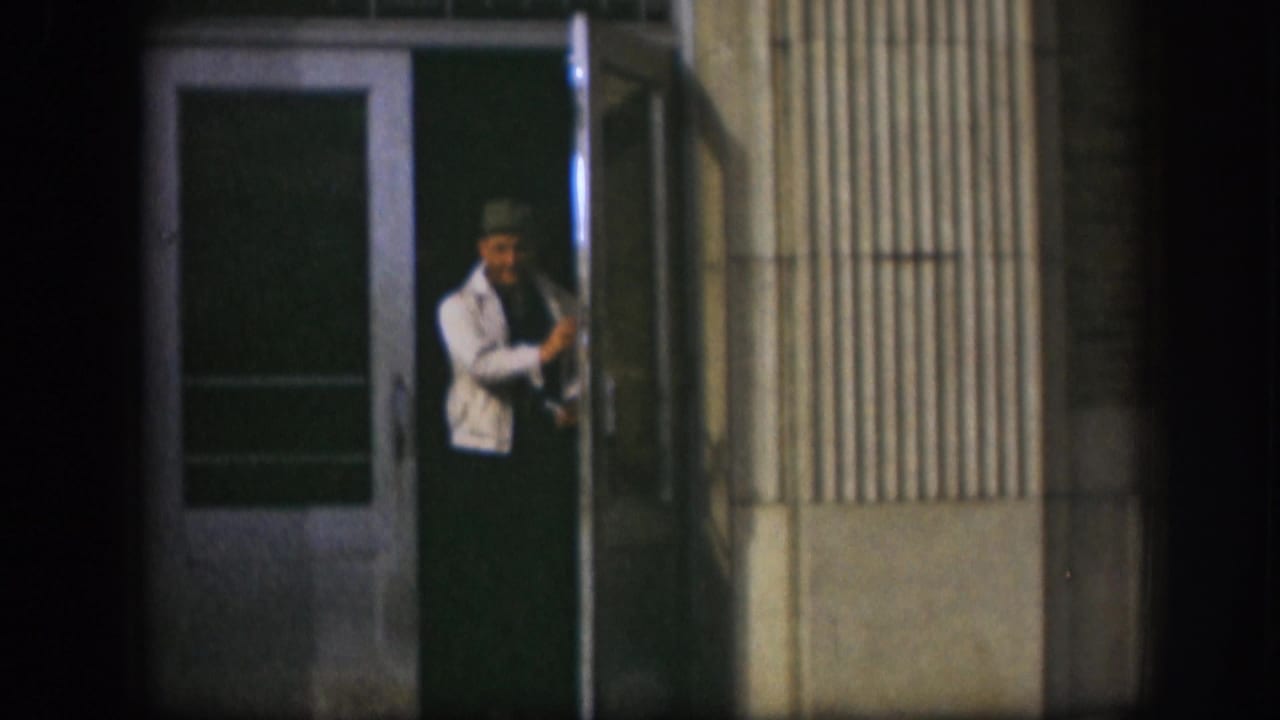 1958 Kansas frame showing a man in a white sweater and hat at a building doorway beside a ribbed exterior wall.