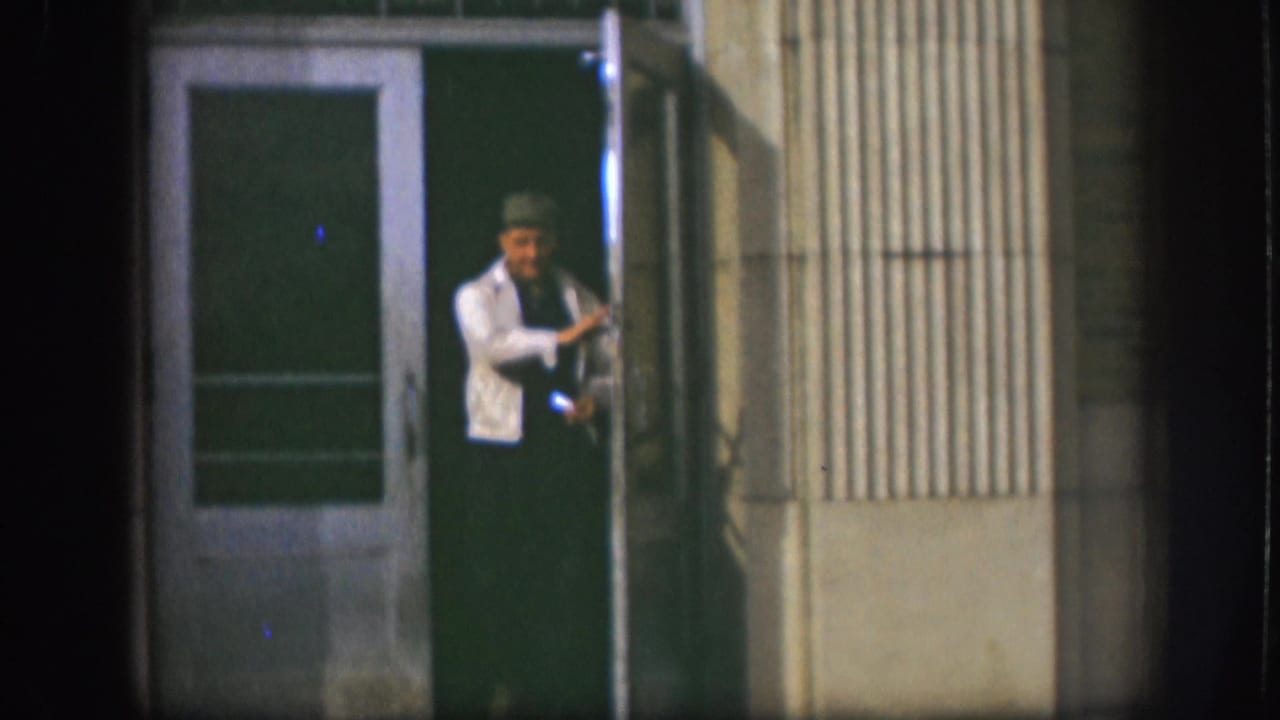In 1958 Kansas home-movie footage, a man in a light jacket and hat stands at a building doorway at night.