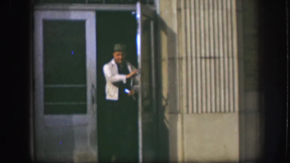 In 1958 Kansas home-movie footage, a man in a light jacket and hat stands at a building doorway at night.