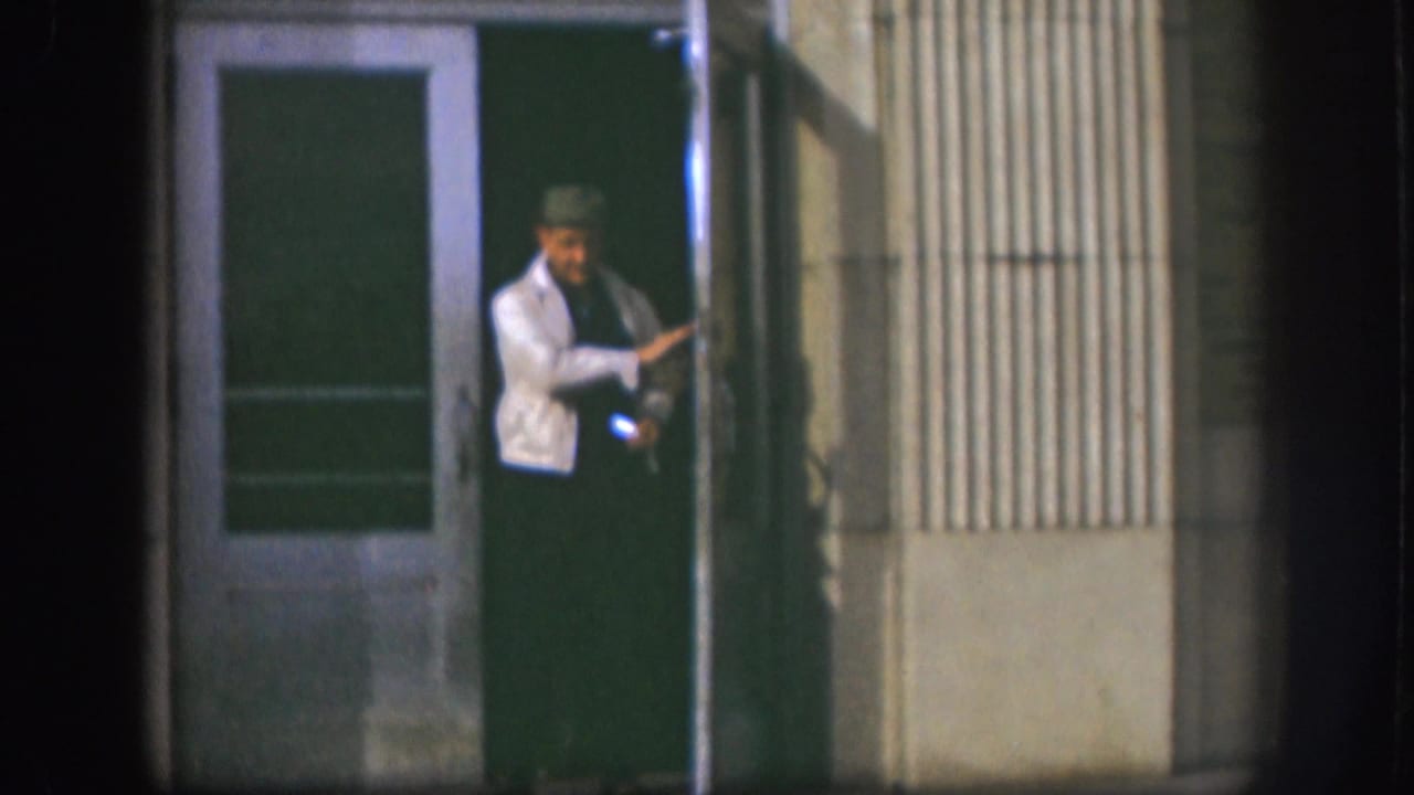 A man in a light jacket and hat stands in a Kansas building doorway, with soft 8mm focus and dark night exposure.