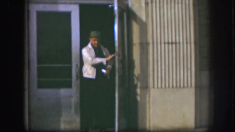 A man in a light jacket and hat stands in a Kansas building doorway, with soft 8mm focus and dark night exposure.
