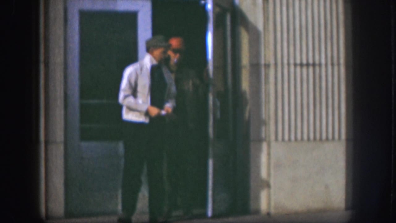 Two adult men stand in the entrance of a Kansas building at night, with one man in a light jacket and another in a red cap.