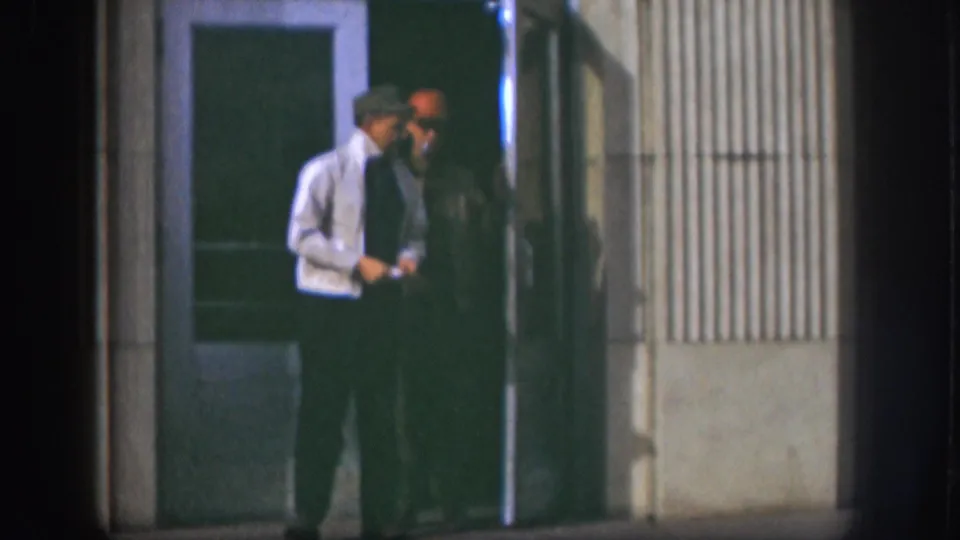 Two adult men stand in the entrance of a Kansas building at night, with one man in a light jacket and another in a red cap.