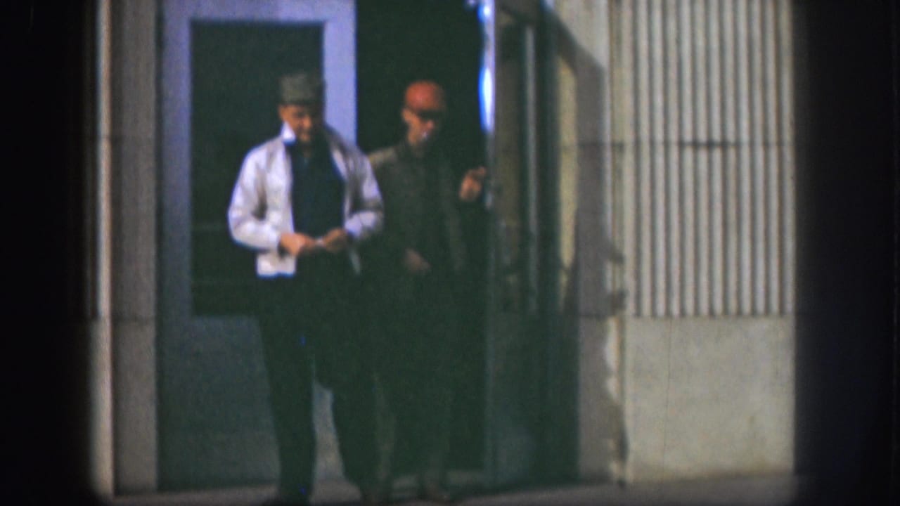 In 1958 Kansas footage, two adult men step away from a building entrance, with the doorway and siding visible behind them.