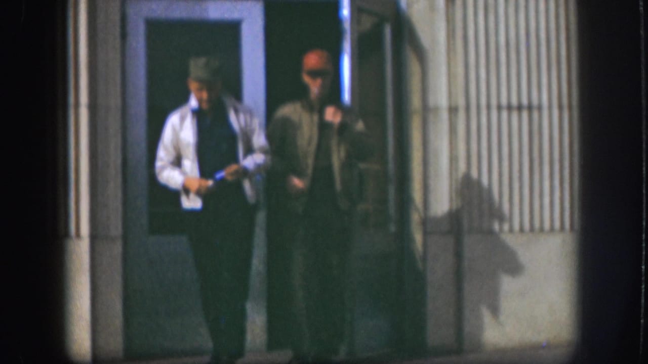 Two adult men walk side by side outside a Kansas doorway in low-light 1958 home-movie footage.