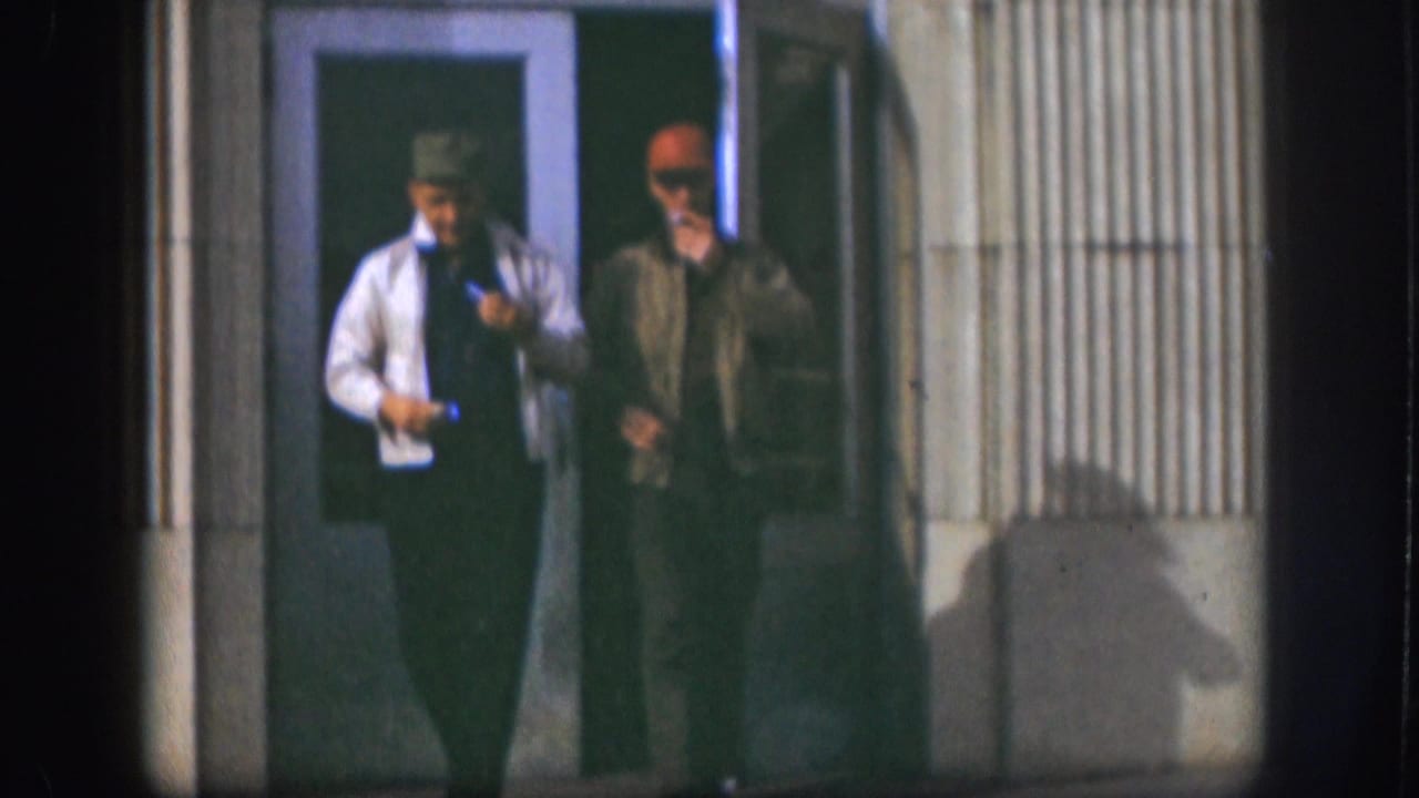 Two men in caps stand outside a doorway, both holding cigarettes.