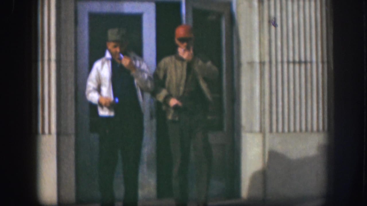 Two adult men in caps and jackets walk from a Kansas building entrance in 1958, with cigarettes visible in their hands.