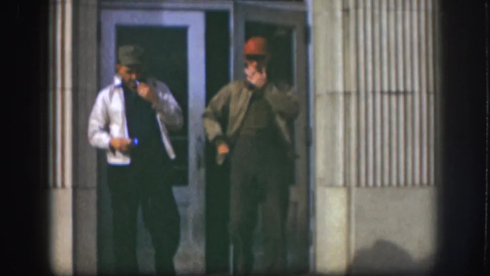 The 1958 Kansas frame shows two adult men smoking beside a doorway, one in a light jacket and one in a brown jacket.