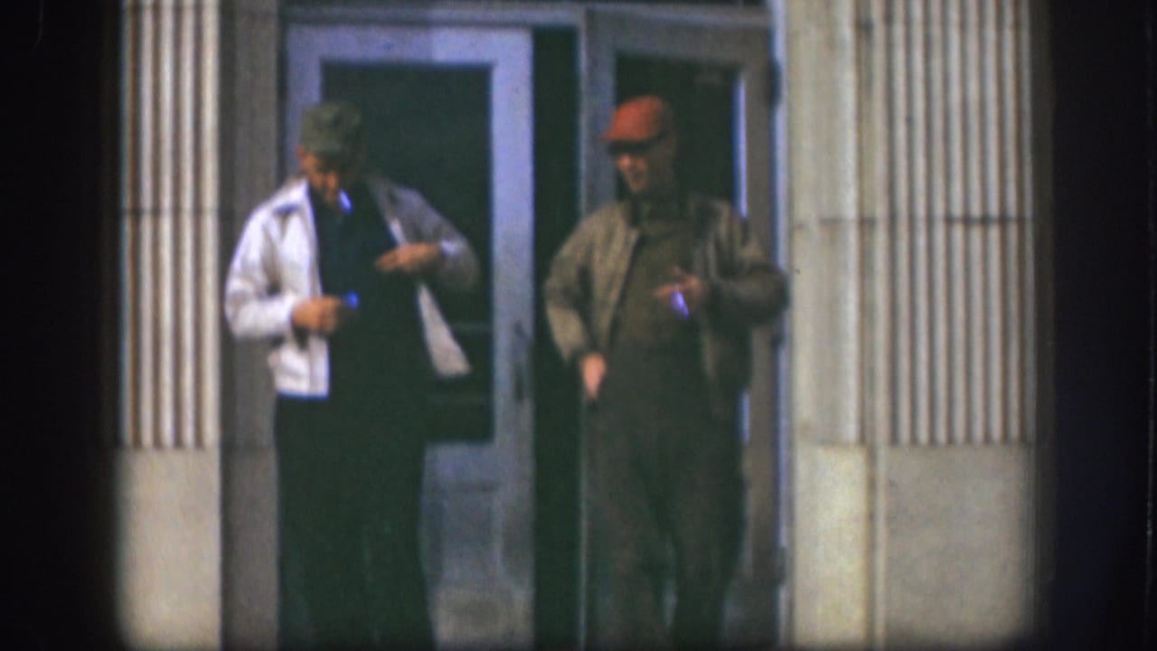 Two adult men in caps and jackets stand outside a Kansas doorway in 1958, with cigarettes and strong wall shadows visible.