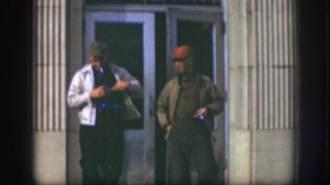 Two adult men in hats stand outside glass doors, one adjusting a white jacket while the other holds a cigarette.