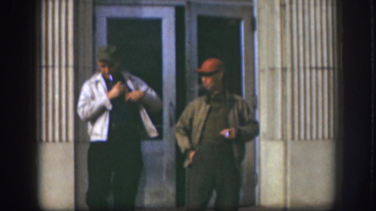 Two adult men in 1950s attire stand outside a Kansas building entrance, framed by glass doors and fluted columns.