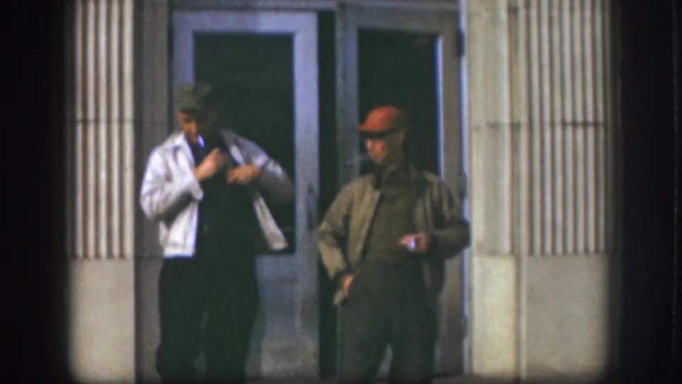 Two adult men in 1950s attire stand outside a Kansas building entrance, framed by glass doors and fluted columns.