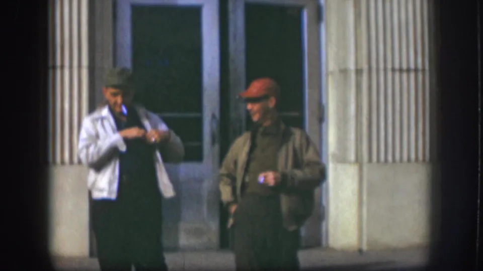 The 1958 Kansas footage shows two adult men outside glass doors, one adjusting his jacket as the other stands with a cigarette.