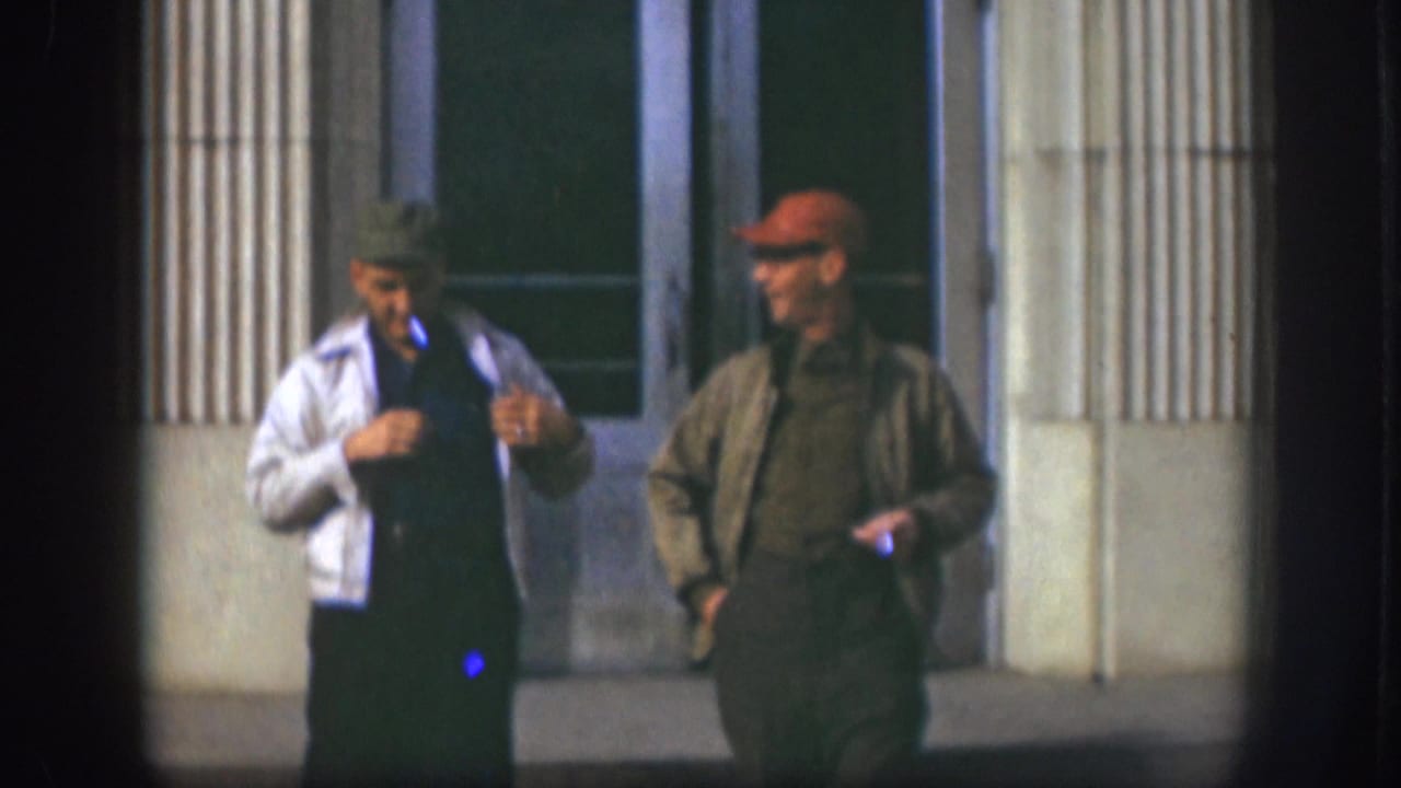 Two adult men in 1950s Kansas stand outside a columned building entrance, one in a white jacket and one in an olive jacket.