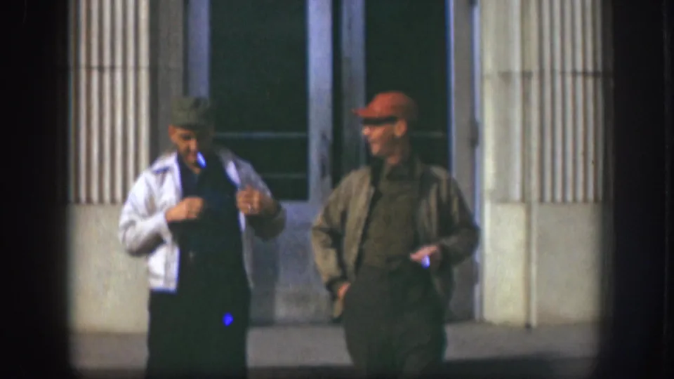 Two adult men in 1950s Kansas stand outside a columned building entrance, one in a white jacket and one in an olive jacket.