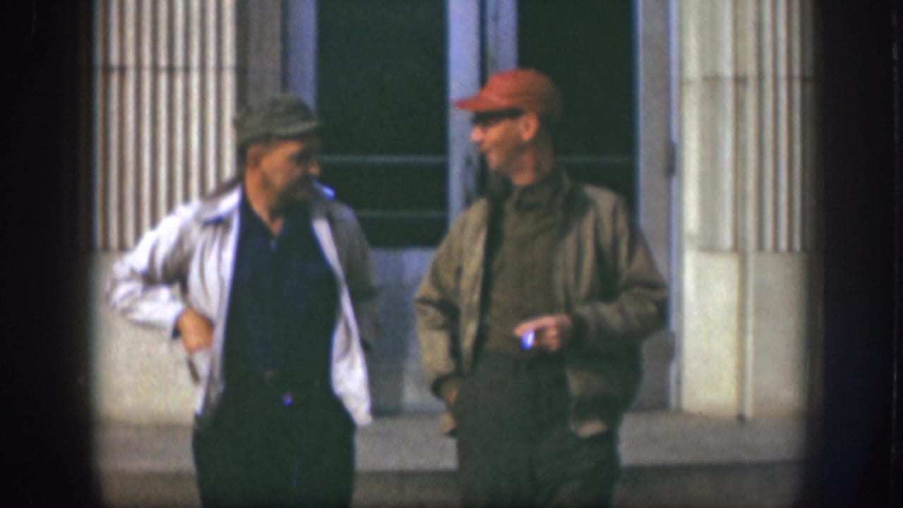Two adult men in caps stand side by side outside a Kansas building, with one smoking during the short break.