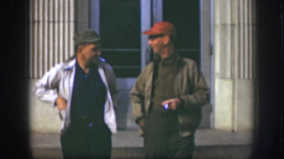 Two adult men in caps stand side by side outside a Kansas building, with one smoking during the short break.