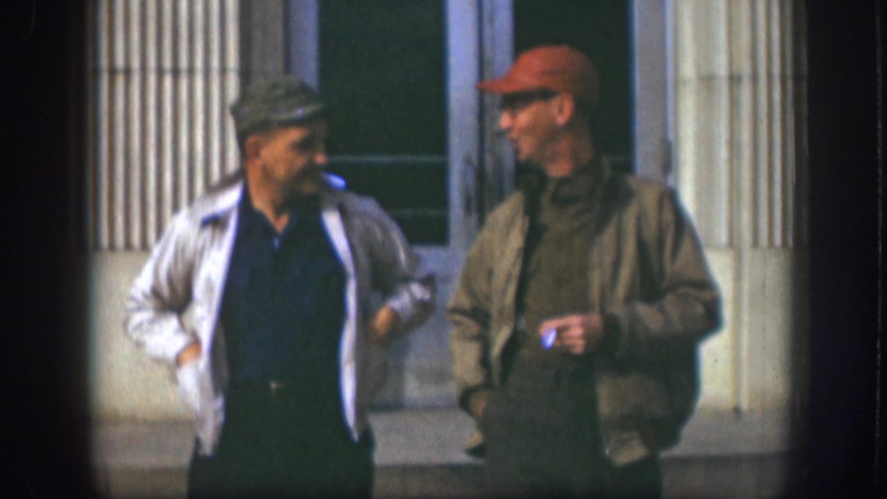 In supported 1958 Kansas footage, two adult men in caps walk and talk outside a building with vertical columns.