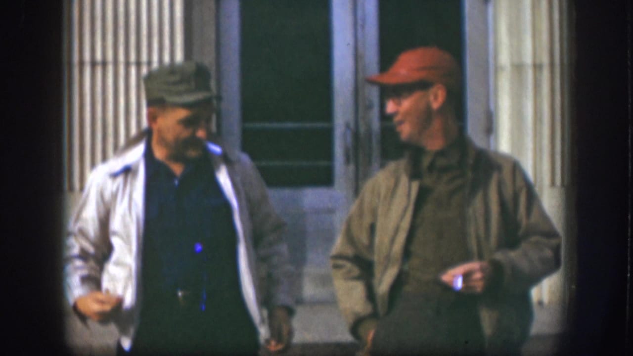 Two adult men in caps converse while moving past a columned exterior in the Kansas home-movie clip.