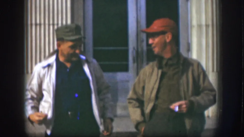 Two adult men in caps converse while moving past a columned exterior in the Kansas home-movie clip.