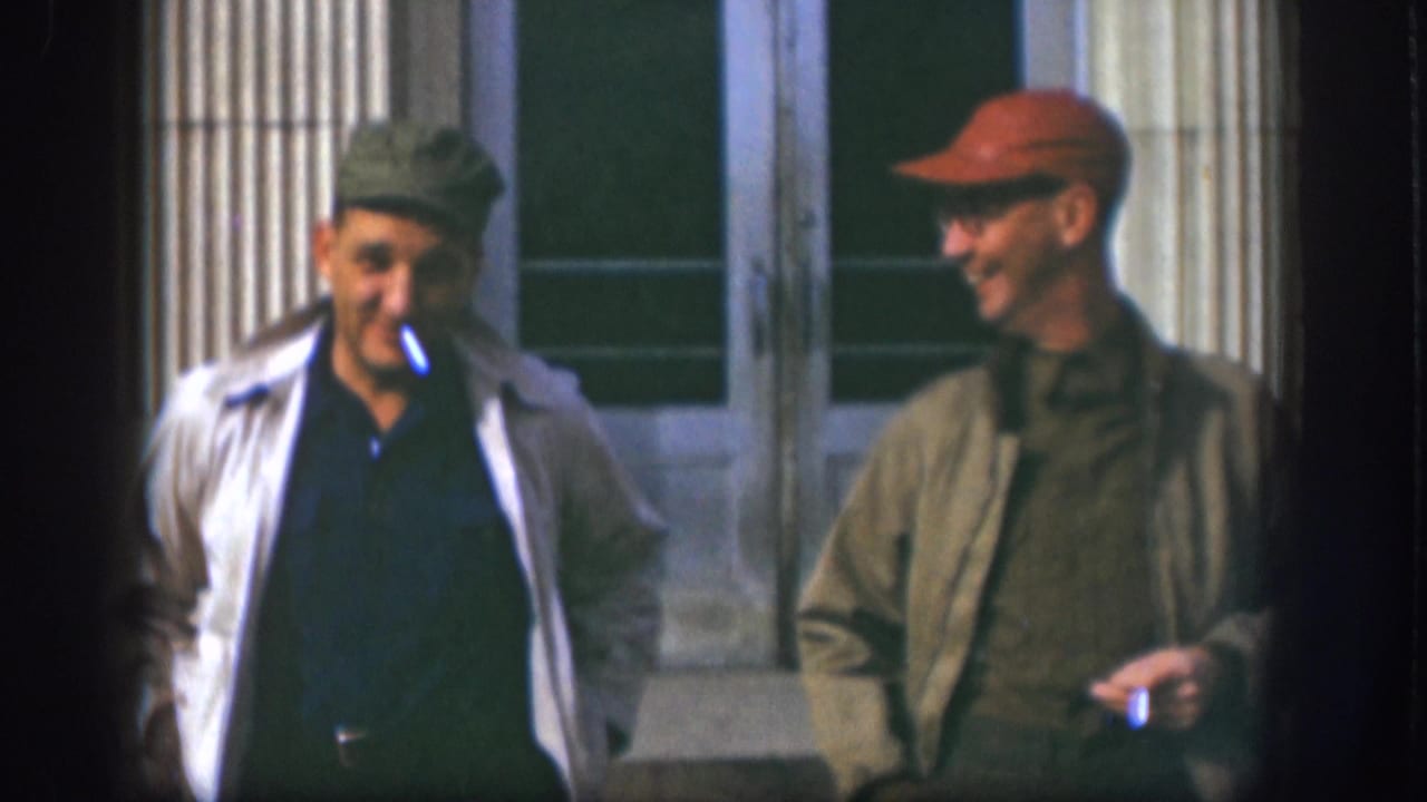 The man in a green cap smokes while walking beside a man in a red cap outside the columned building.